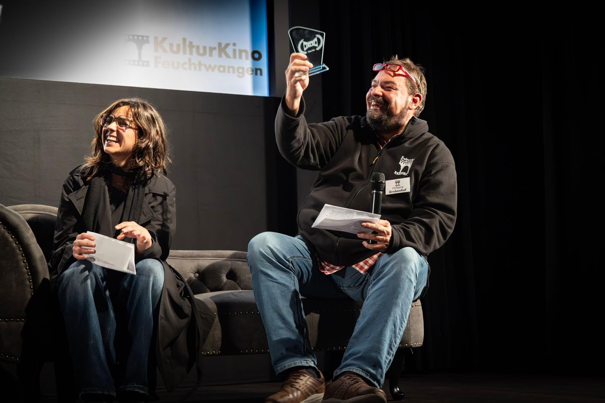 Preisverleihung im KulturKino Feuchtwangen, Moderator hält lachend die Trophäe hoch
